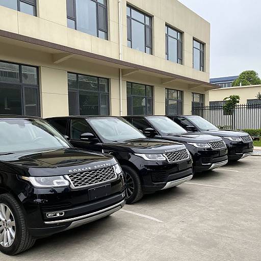 Photograph of four shiny black SUVs parked in front of a modern beige building with large windows, under a cloudy sky.