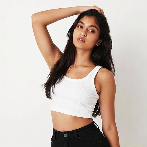 Photograph of a young woman with long black hair, wearing a white ribbed crop top and black high-waisted pants, posing with one arm