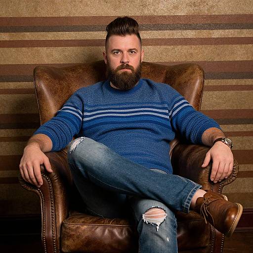 Photograph of a bearded man with a full head of dark hair, wearing a blue striped sweater and ripped jeans, sitting in a brown leather arm