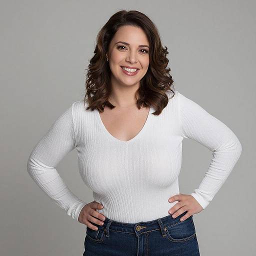 Photograph of a smiling woman with dark brown, wavy hair, wearing a white ribbed long-sleeve top and blue jeans, standing against