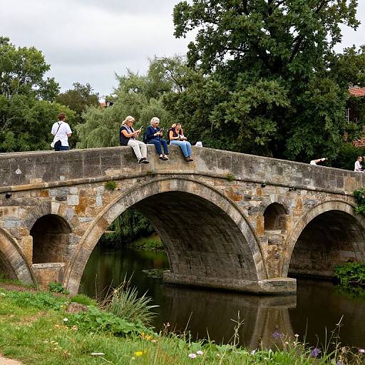 Photograph of six people, including children and adults, sitting on a rustic stone bridge with three arches, surrounded by lush green trees and a grass