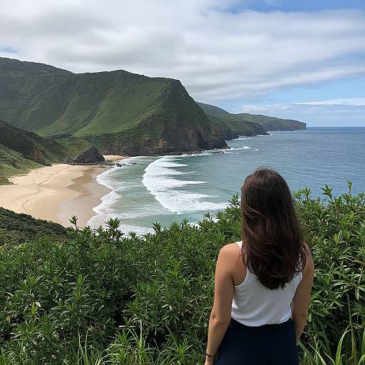 Photograph of a woman with long brown hair in a white tank top and black skirt, standing on a grassy cliff, overlooking a stunning coastal landscape