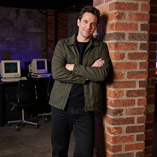 Photograph of a smiling man with short dark hair, black shirt, and green jacket, standing with arms crossed against a brick pillar in a dimly
