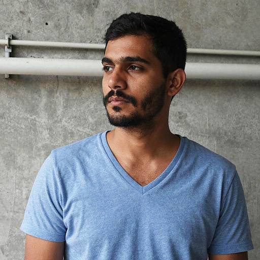 Photograph of a bearded South Asian man with short black hair, wearing a light blue V-neck t-shirt, standing against a textured gray concrete wall