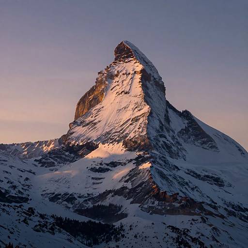 Photograph of a snow-covered mountain peak bathed in warm sunset light, with rugged textures and a clear, purple-tinged sky backdrop.