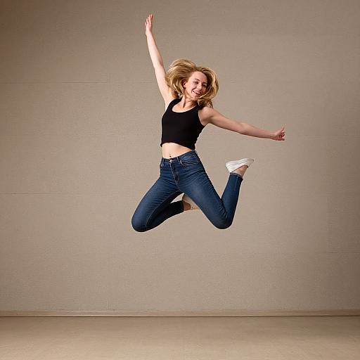 Photograph of a blonde woman with a fair complexion, wearing a black tank top and blue jeans, mid-air jumping against a plain beige wall and floor