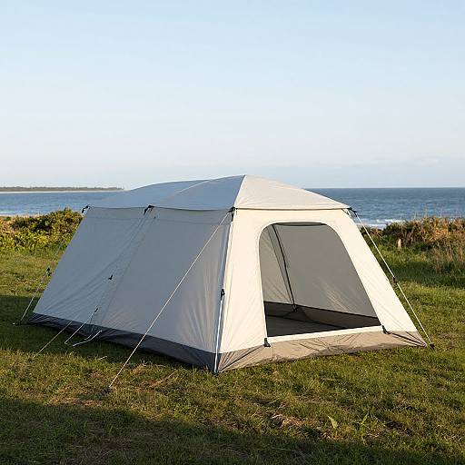 Photograph of a white and gray camping tent with black accents, set on grassy terrain near a serene ocean view under clear blue sky.
