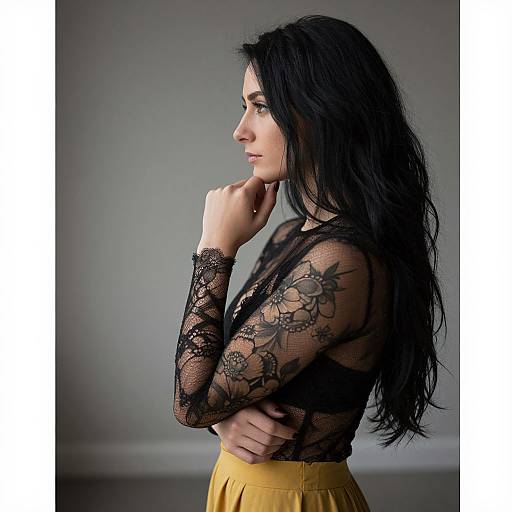 Photograph of a pensive woman with long black hair, wearing a sheer black lace top and yellow skirt, against a gray background. She rests her