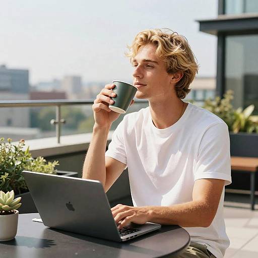 Photograph of a blond, fair-skinned man in a white t-shirt, sipping from a green mug while working on a laptop on a sun