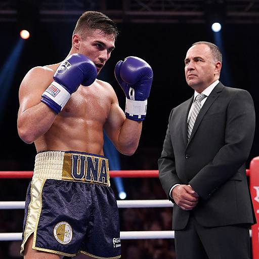 Photograph of a muscular, shirtless male boxer in navy and gold trunks, blue gloves, and focused expression, standing in a boxing ring with