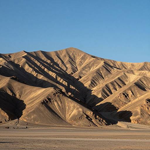 Photograph of a desert mountain range with golden sunlight casting dramatic shadows on rugged, brown hills under a clear, bright blue sky.