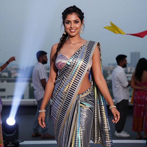 Photograph of a smiling Indian woman with long braided hair, wearing a shimmering silver and black striped saree, on a rooftop with cityscape