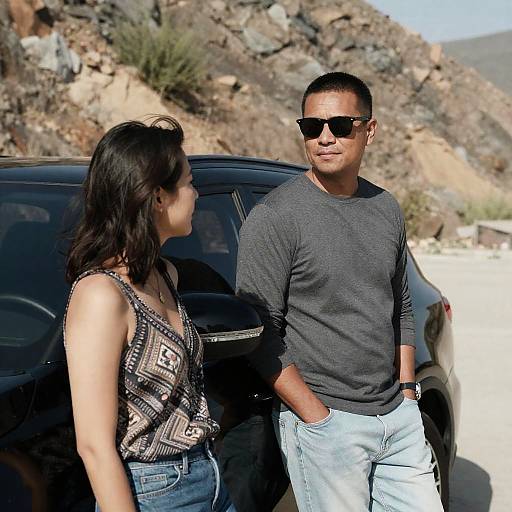 Photograph of an Asian couple standing beside a black car in a rocky desert landscape; woman in patterned top, man in gray shirt and jeans,