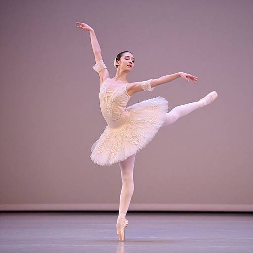 Photograph of a graceful ballerina performing a pirouette, wearing a white tutu and ballet slippers, against a gray stage backdrop.