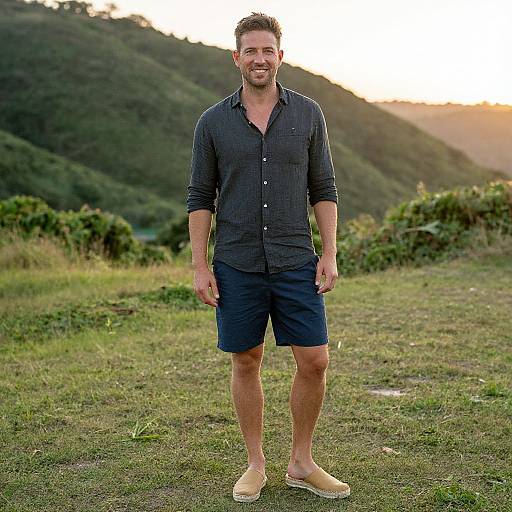 Photograph of a smiling, bearded man in a dark checkered shirt, navy shorts, and beige slip-on shoes, standing on grassy terrain