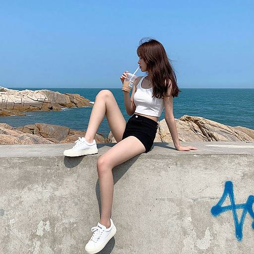 Photograph of a young woman with long brown hair, white tank top, black shorts, and white sneakers, sipping a drink on a concrete seaw