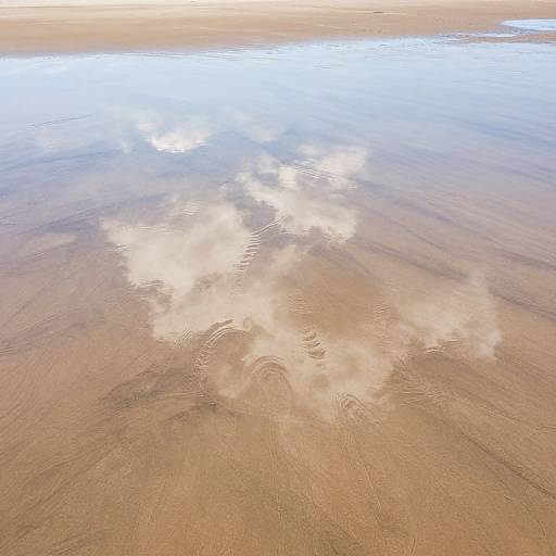 Aerial photograph of a sandy beach with gentle ripples reflecting white, fluffy clouds on a calm, shallow shore.