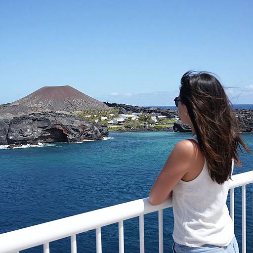 Photograph of a woman with long dark hair in a white tank top, standing on a white railing, gazing at a volcanic island and clear blue