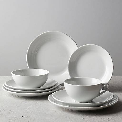 Photograph of white ceramic teacup and saucer set with two large plates, arranged on a gray surface, illuminated by soft light.