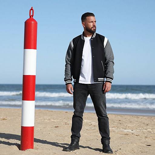 Man in Varsity Jacket Standing on Beach Near Red and White Pole