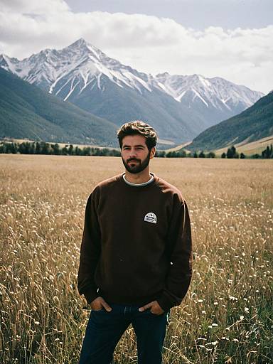 Man in Brown Sweatshirt Standing in Field with Snowy Mountains Background