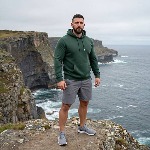 Man Standing on Rocky Cliff Overlooking Ocean in Green Hoodie and Gray Shorts